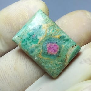 Könnte beinhalten: Ein rechteckiger Edelstein mit einer leuchtend grünen Grundfarbe, gesprenkelt mit Gelb und Braun. Der Stein weist ein zentrales, auffälliges rosa-rotes Quadrat auf. Der Edelstein wird von einer behandschuhten Hand gehalten, die seine natürliche Schönheit zeigt.