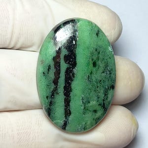 Könnte beinhalten: Ovaler Edelstein mit überwiegend grüner Farbe und auffälligen schwarzen Adern. Die polierte Oberfläche reflektiert das Licht und zeigt die natürlichen Muster. Der Stein wird von einer behandschuhten Hand gehalten, was seine Größe und Form hervorhebt.