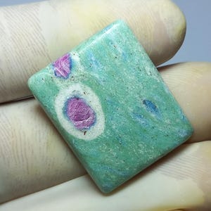 Könnte beinhalten: Ein quadratischer Edelstein mit hellgrüner Grundfarbe und rosa und blauen Einschlüssen. Der polierte Stein hat ein einzigartiges Muster mit einem prominenten rosa Zentrum, das von einem weißen und blauen Ring umgeben ist.