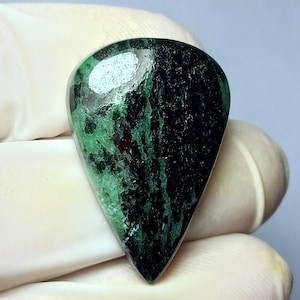 May include: A teardrop-shaped gemstone with a polished surface. The stone features a mix of dark green and black colors with streaks and patches. The gemstone is held by a gloved hand, showcasing its natural beauty and unique pattern.