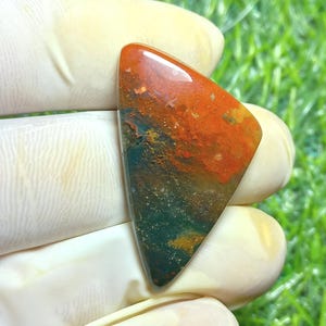 Puede incluir: Una piedra preciosa pulida con forma triangular. La piedra presenta una mezcla de colores, incluyendo tonos rojo intenso, naranja y verde. La superficie es lisa y reflectante, mostrando los patrones y texturas naturales de la piedra.