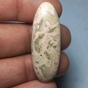 Può includere: Una gemma ovale con una base color crema, con motivi a spirale rosa e beige. La superficie lucida mostra un design naturale e organico, con aree più scure e macchiate. La pietra è tenuta in mano, evidenziando le sue dimensioni e la sua forma. Misura circa 5 cm.