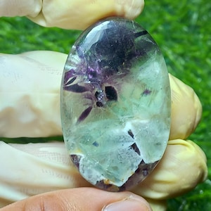 May include: An oval-shaped, polished fluorite gemstone. The stone features a blend of colors, including deep purple, clear, and light green. The gemstone is held by a gloved hand, with a green background.