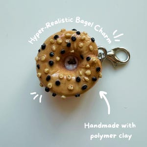 Pode incluir: Um charm de bagel hiper-realista feito com argila polimérica. O charm é bege com detalhes em marrom escuro e marrom claro, e um fecho prateado. O texto "Hyper-Realistic Bagel Charm" e "Handmade with polymer clay" são visíveis.
