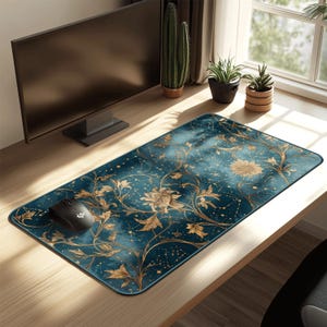 Puede incluir: Alfombrilla de ratón rectangular de color turquesa con un diseño floral y de constelaciones en dorado. Un ratón de ordenador negro está sobre la alfombrilla. La alfombrilla está sobre un escritorio de madera cerca de un monitor de ordenador y plantas.