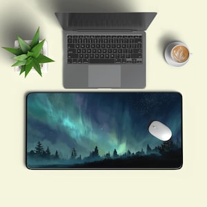 Puede incluir: Alfombrilla de ratón rectangular con un diseño de paisaje que representa un bosque bajo la aurora boreal. También se ven un portátil, un ratón blanco, una pequeña planta en maceta y una taza de café.