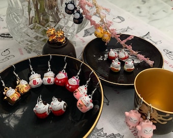 May include: A collection of Maneki Neko cat earrings in various colors, including gold, red, white, and pink. The earrings are displayed on black plates and a gold-colored cup, with a floral arrangement in the background. The earrings are a charming accessory.