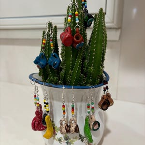 May include: A collection of handmade earrings in various shapes and colors, including red, blue, yellow, and green, are displayed on a cactus plant. The earrings are adorned with colorful beaded accents and hang from silver hooks. The cactus is in a white and blue floral pot.