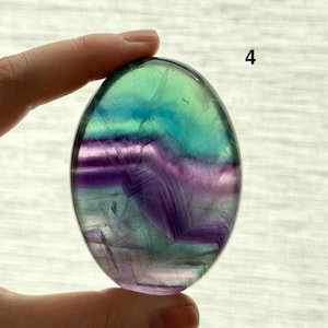 Puede incluir: Una piedra de fluorita pulida, de forma ovalada. La piedra presenta capas de tonos morados, azules y verdes. El n&uacute;mero "4" es visible en la esquina superior derecha. La piedra se sostiene entre dos dedos.