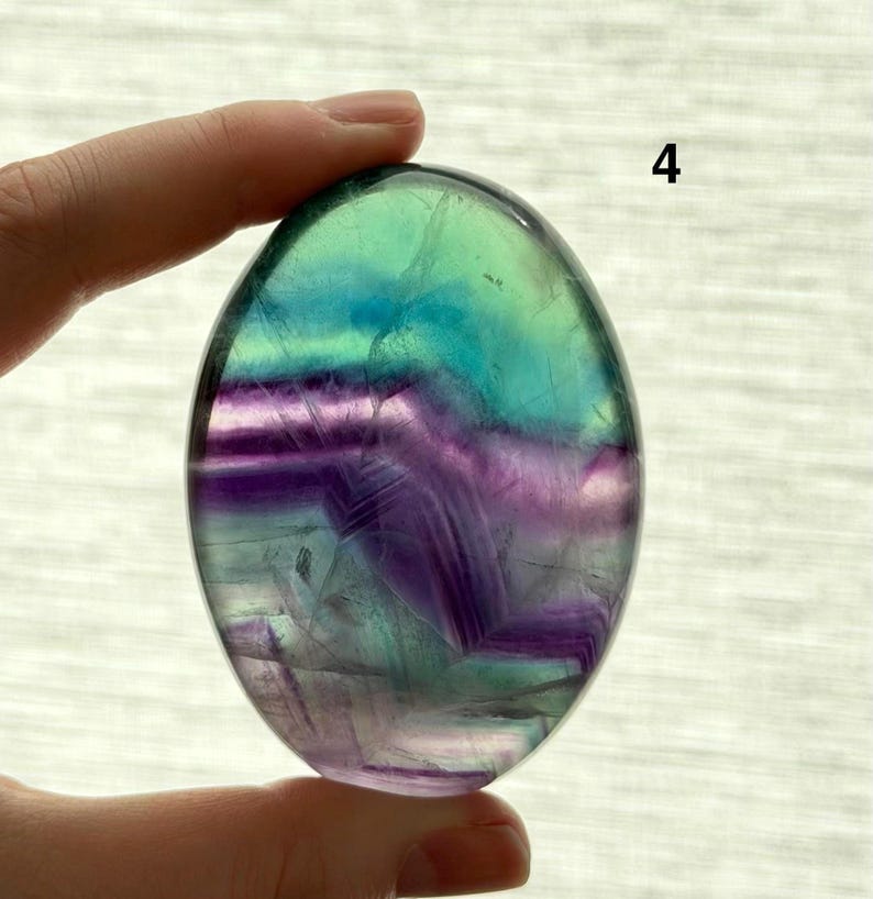 Puede incluir: Una piedra de fluorita pulida, de forma ovalada. La piedra presenta capas de tonos morados, azules y verdes. El n&uacute;mero "4" es visible en la esquina superior derecha. La piedra se sostiene entre dos dedos.