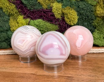 May include: Three polished gemstone spheres in shades of pink, white, and gray, each displayed on a clear acrylic stand. The spheres have unique banding patterns. The background features a textured moss wall.