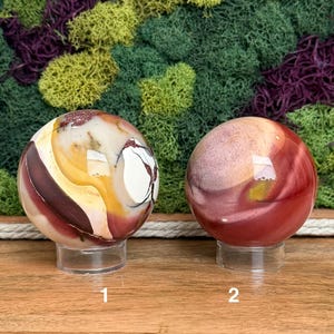 Puede incluir: Dos piedras esf&eacute;ricas pulidas sobre soportes circulares transparentes. La esfera izquierda presenta patrones de remolinos en crema, amarillo y granate oscuro con vetas. La esfera derecha muestra un degradado de tonos rojo, rosa y beige apagados.