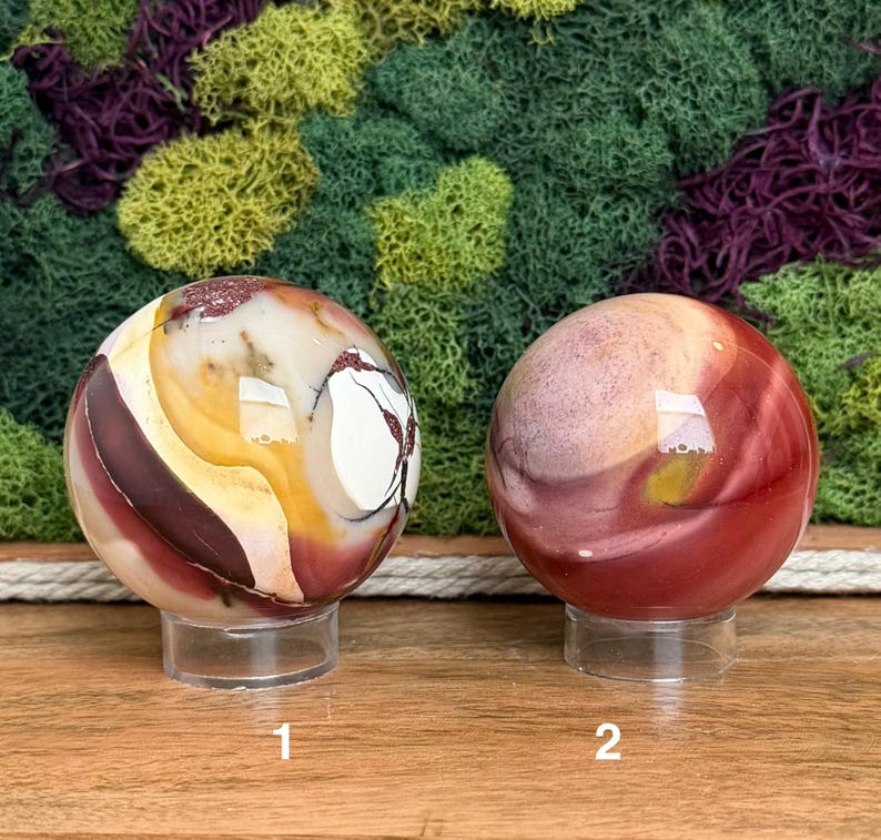 Puede incluir: Dos piedras esf&eacute;ricas pulidas sobre soportes circulares transparentes. La esfera izquierda presenta patrones de remolinos en crema, amarillo y granate oscuro con vetas. La esfera derecha muestra un degradado de tonos rojo, rosa y beige apagados.