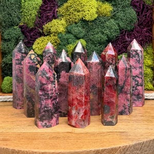 Puede incluir: Una colecci&oacute;n de torres de piedras preciosas rosas y negras con puntas. Los cristales est&aacute;n dispuestos sobre una superficie de madera, con un tel&oacute;n de fondo de musgo verde, morado y amarillo.