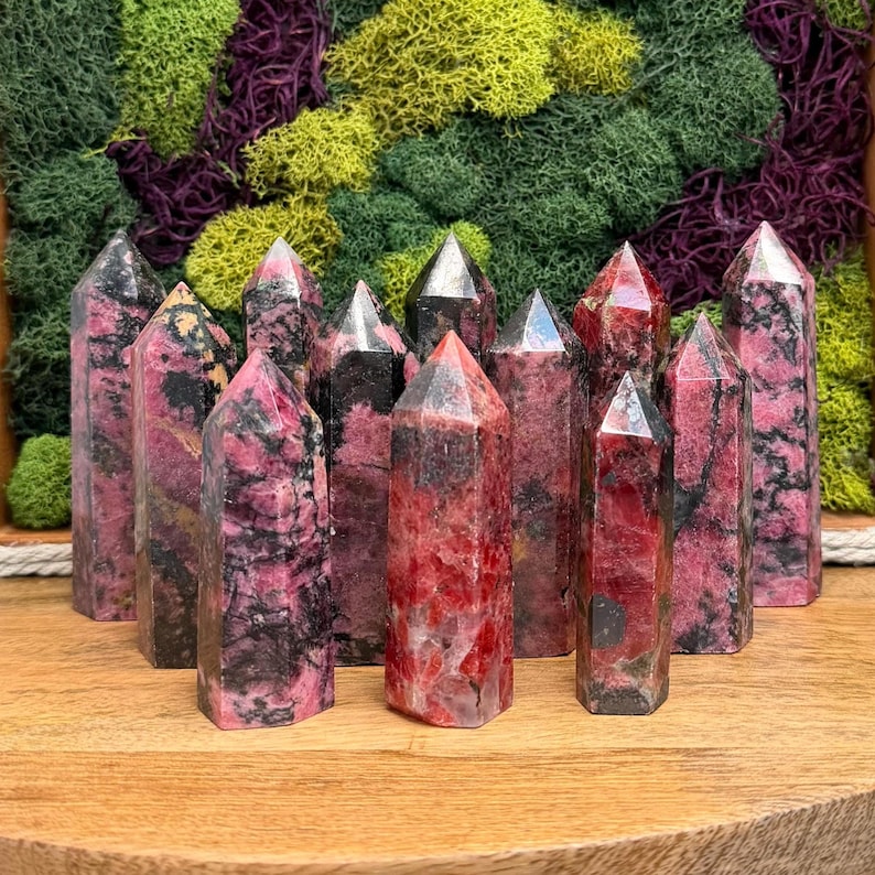 Puede incluir: Una colecci&oacute;n de torres de piedras preciosas rosas y negras con puntas. Los cristales est&aacute;n dispuestos sobre una superficie de madera, con un tel&oacute;n de fondo de musgo verde, morado y amarillo.
