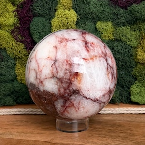 May include: A polished, spherical gemstone with a white base and intricate red and brown veining. The sphere sits on a clear, circular stand, showcasing its natural patterns. The background features a mix of green and burgundy moss.