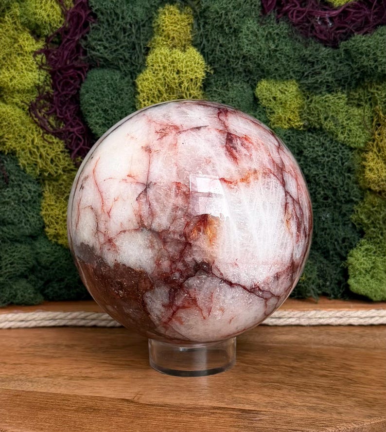 May include: A polished, spherical gemstone with a white base and intricate red and brown veining. The sphere sits on a clear, circular stand, showcasing its natural patterns. The background features a mix of green and burgundy moss.