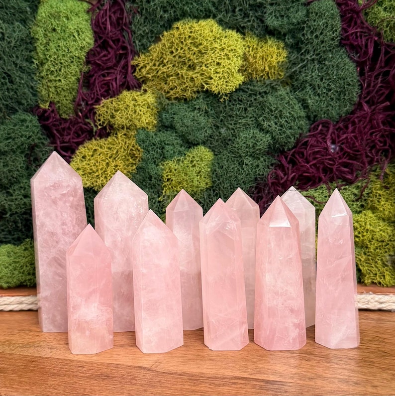 May include: A collection of ten rose quartz crystal points, each featuring a polished, multi-faceted hexagonal shape with a pointed top. The stones exhibit a translucent, pale pink hue with natural internal fractures and varying heights.