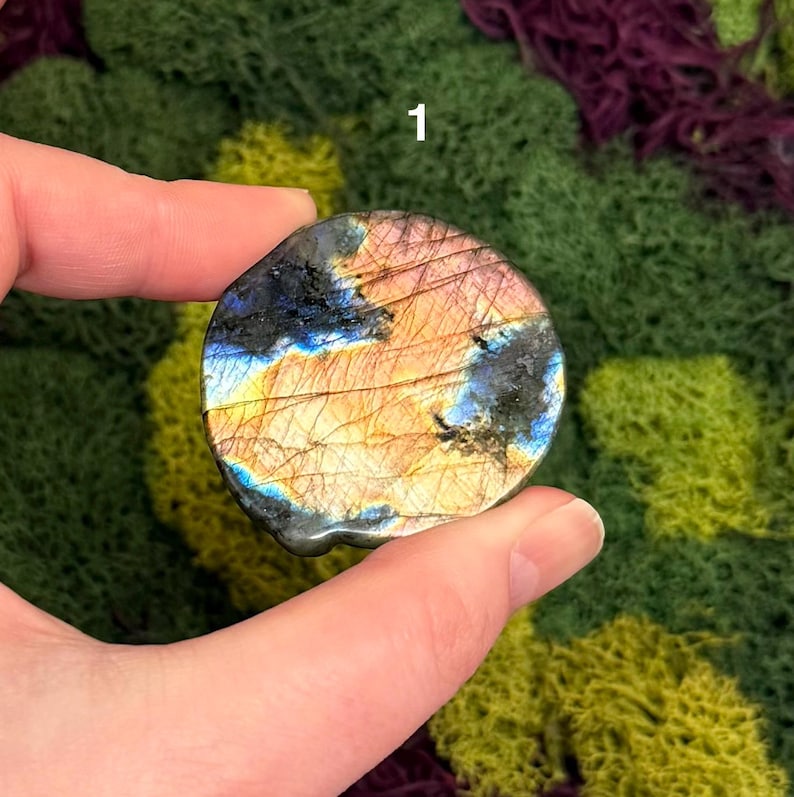 May include: A round, polished gemstone with a mix of colors, including blue, gold, and black. The stone is held between fingers and thumb. The number "1" is visible in the upper right corner.