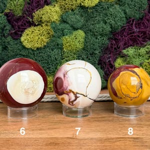 Puede incluir: Tres esferas de piedra pulida sobre soportes circulares transparentes. La esfera izquierda es de color rojo oscuro con un gran centro blanco. La central es blanca con vetas granates. La derecha es amarilla con patrones rojos y marrones.