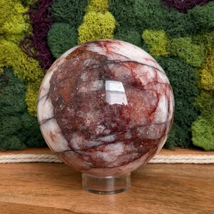 May include: A polished sphere of red and white stone, resting on a clear stand. The orb's surface displays intricate patterns of red veining against a white background. The sphere is set against a backdrop of green and purple moss.