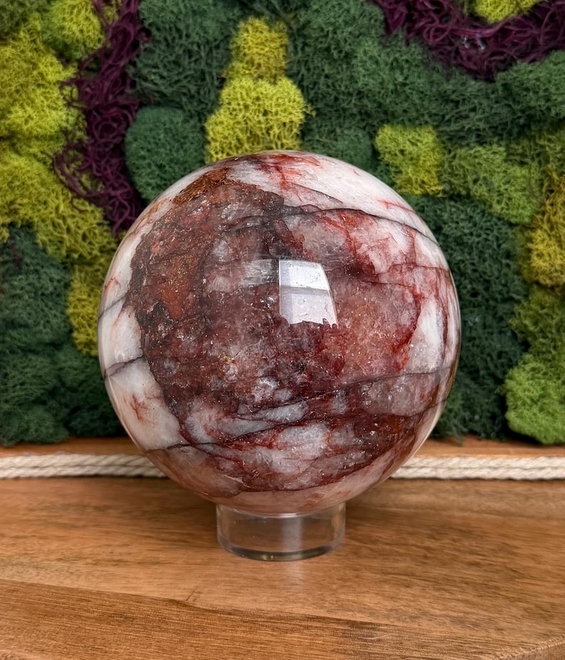 May include: A polished sphere of red and white stone, resting on a clear stand. The orb's surface displays intricate patterns of red veining against a white background. The sphere is set against a backdrop of green and purple moss.