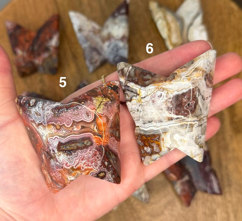 May include: A collection of butterfly-shaped agate stones in shades of brown, red, white, and cream. The stones have unique patterns and textures, with visible numbering. The stones are held in a hand, showcasing their natural beauty.