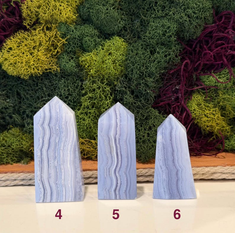 May include: Three blue lace agate obelisks with white veining, numbered 4, 5, and 6. The obelisks are displayed against a backdrop of green, yellow, and burgundy moss.