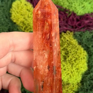 May include: A tall, reddish-orange crystal point with a faceted surface. The crystal has a pointed top and is held in a hand. The background features green and purple textured elements. The number "8" is visible in the background.