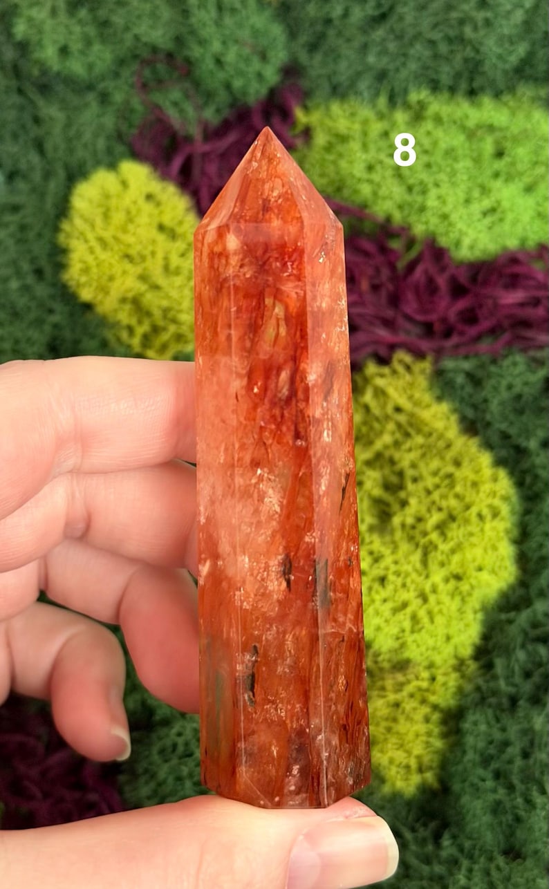 May include: A tall, reddish-orange crystal point with a faceted surface. The crystal has a pointed top and is held in a hand. The background features green and purple textured elements. The number "8" is visible in the background.