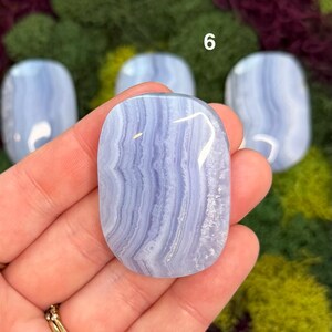 May include: A polished, oval-shaped blue lace agate stone with white and light blue banding. The stone is held in a hand, with other similar stones visible in the background. The number 6 is visible in the background.