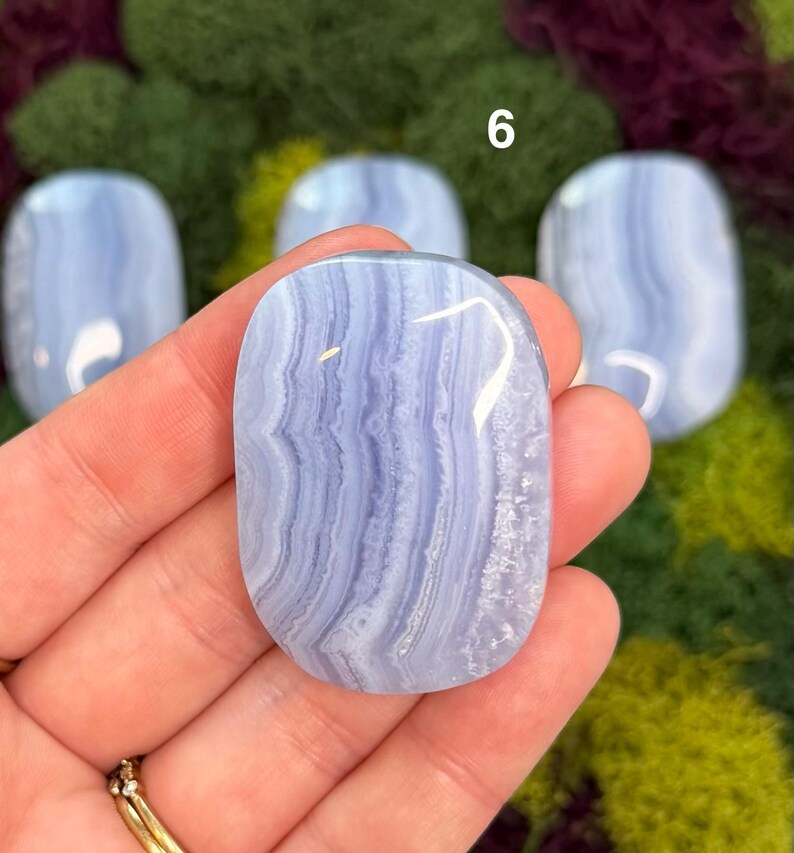 May include: A polished, oval-shaped blue lace agate stone with white and light blue banding. The stone is held in a hand, with other similar stones visible in the background. The number 6 is visible in the background.
