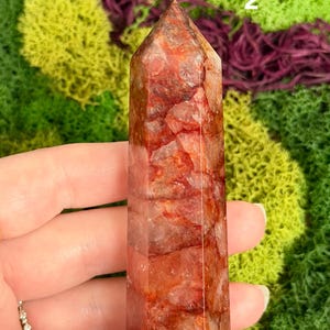 May include: A reddish-brown quartz crystal tower with a pointed top. The crystal is held in a hand and has a natural, textured appearance. The background features green and purple moss.