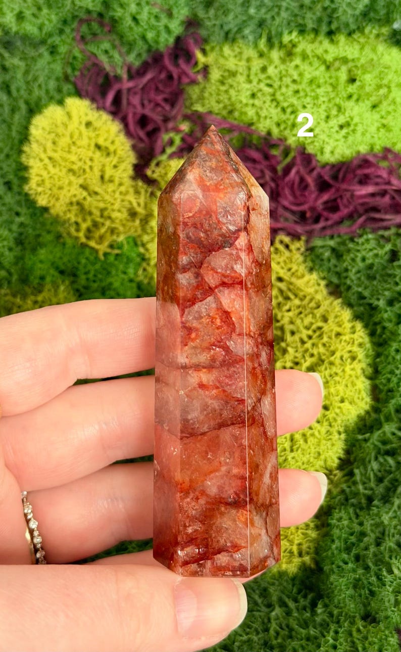 May include: A reddish-brown quartz crystal tower with a pointed top. The crystal is held in a hand and has a natural, textured appearance. The background features green and purple moss.