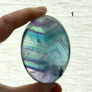 Puede incluir: Una piedra de fluorita ovalada pulida, con una mezcla de tonos azules, verdes y morados. La piedra transl&uacute;cida muestra patrones internos y el n&uacute;mero "1" es visible en la esquina superior derecha.