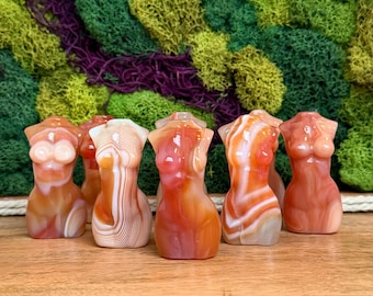 Puede incluir: Una colección de seis figuras de piedra pulida talladas con forma de torso humano. Cada escultura presenta patrones de bandas distintos en tonos naranja, blanco y marrón rojizo, mostrando las variaciones naturales del material de ágata.