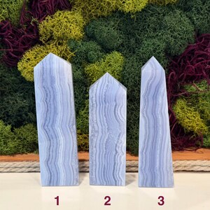 May include: Three blue lace agate obelisks with a layered, banded pattern. The obelisks are numbered 1, 2, and 3. The background features a mix of green, yellow, and burgundy moss.
