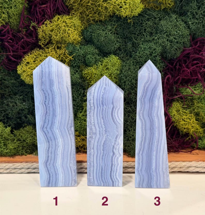 May include: Three blue lace agate obelisks with a layered, banded pattern. The obelisks are numbered 1, 2, and 3. The background features a mix of green, yellow, and burgundy moss.