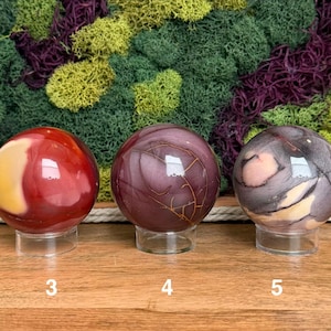 Puede incluir: Tres esferas de piedra pulida sobre soportes circulares transparentes. La primera esfera es roja y amarilla, la segunda es de color p&uacute;rpura intenso con finas vetas doradas, y la tercera es una mezcla de gris, p&uacute;rpura y crema con vetas.