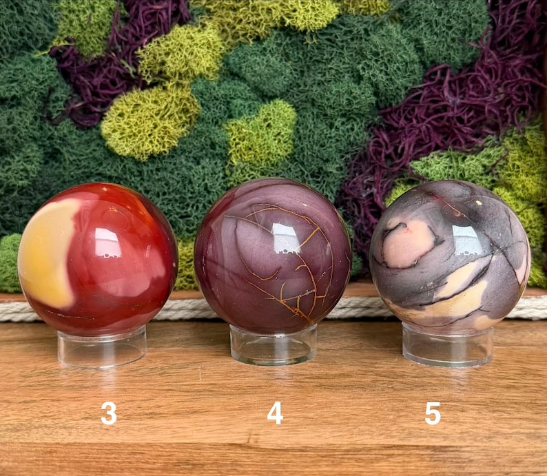 Puede incluir: Tres esferas de piedra pulida sobre soportes circulares transparentes. La primera esfera es roja y amarilla, la segunda es de color p&uacute;rpura intenso con finas vetas doradas, y la tercera es una mezcla de gris, p&uacute;rpura y crema con vetas.