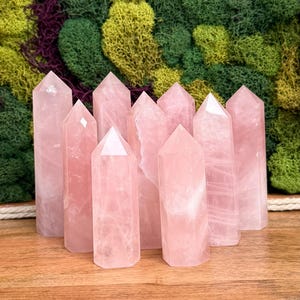 Puede incluir: Un conjunto de nueve torres de cristal de cuarzo rosa, cada una con una forma hexagonal pulida y una punta superior. Las piedras muestran diversos tonos de rosa pálido translúcido, exhibiendo fracturas internas naturales y texturas cristalinas.