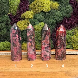Puede incluir: Cuatro torres de cristal de rodonita pulidas con una mezcla de tonos rosa, negro y gris. Cada torre tiene una parte superior puntiaguda y una base rectangular. Los cristales est&aacute;n numerados del 1 al 4.