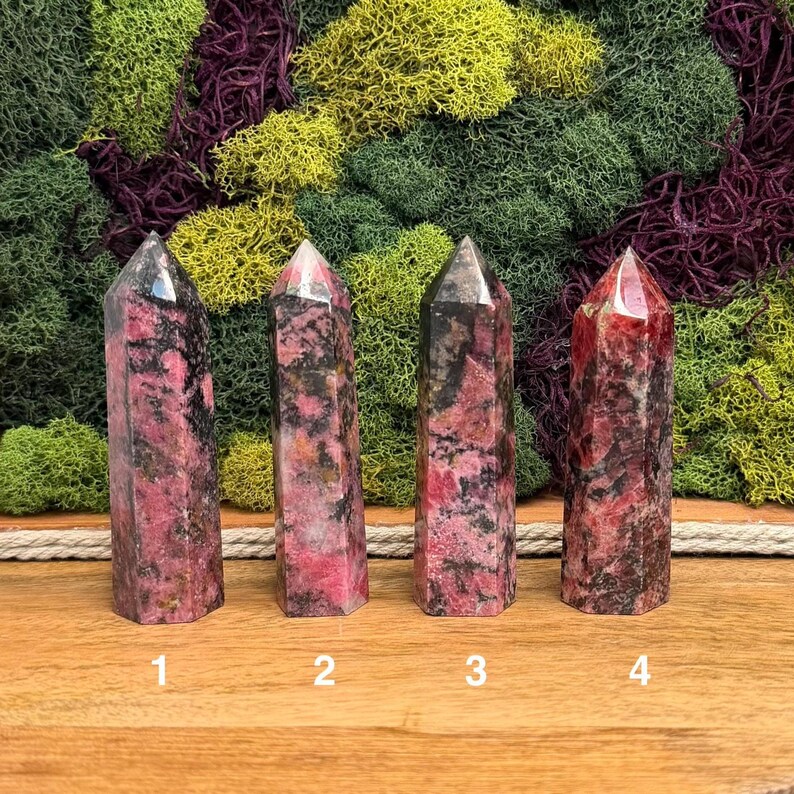 Puede incluir: Cuatro torres de cristal de rodonita pulidas con una mezcla de tonos rosa, negro y gris. Cada torre tiene una parte superior puntiaguda y una base rectangular. Los cristales est&aacute;n numerados del 1 al 4.