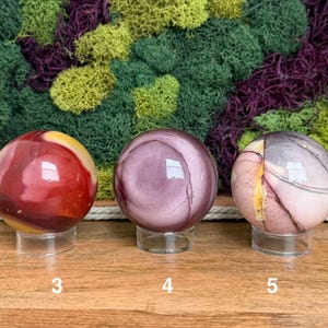Puede incluir: Tres esferas de piedra pulida sobre soportes circulares transparentes, etiquetadas con los n&uacute;meros 3, 4 y 5. La primera esfera es roja y amarilla, la segunda es de color malva s&oacute;lido con patrones circulares y la tercera es rosa p&aacute;lido y gris.