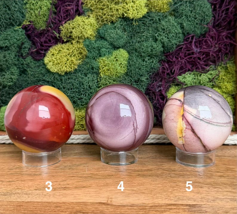 Puede incluir: Tres esferas de piedra pulida sobre soportes circulares transparentes, etiquetadas con los n&uacute;meros 3, 4 y 5. La primera esfera es roja y amarilla, la segunda es de color malva s&oacute;lido con patrones circulares y la tercera es rosa p&aacute;lido y gris.