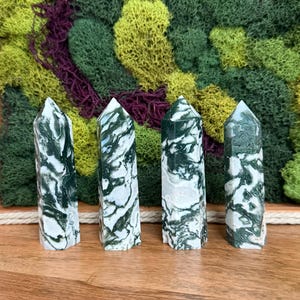May include: Four polished gemstone towers with a green and white marbled pattern. The crystals are standing on a wooden surface, with a backdrop of green, yellow, and purple moss.