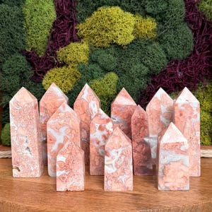 Puede incluir: Una colección de torres de cristal con patrones rosas y blancos con puntas puntiagudas. Los cristales están dispuestos sobre una superficie de madera, con un fondo de musgo verde, amarillo y morado.