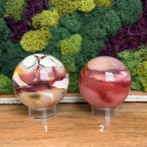 Puede incluir: Dos piedras esf&eacute;ricas pulidas sobre soportes circulares transparentes. La piedra de la izquierda presenta vetas en crema, rojo oscuro y marr&oacute;n oscuro. La piedra de la derecha es una esfera lisa y s&oacute;lida en tonos rosa empolvado y rojo.