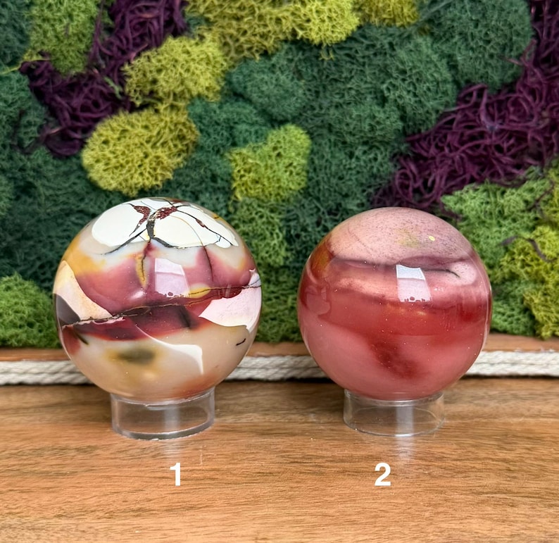 Puede incluir: Dos piedras esf&eacute;ricas pulidas sobre soportes circulares transparentes. La piedra de la izquierda presenta vetas en crema, rojo oscuro y marr&oacute;n oscuro. La piedra de la derecha es una esfera lisa y s&oacute;lida en tonos rosa empolvado y rojo.