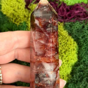 May include: A clear, rectangular crystal tower with red and white inclusions. The crystal has a pointed top and is held in a hand. The number "11" is visible on the crystal. The background is green and purple.
